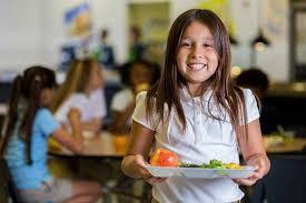 A smiling girl holds a plate of food in a school cafeteria, with other children in the background.